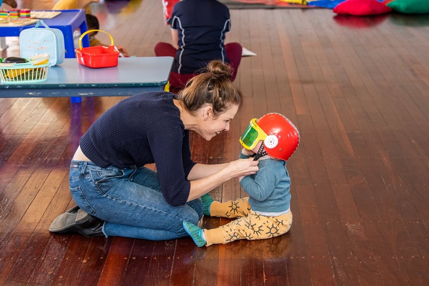 Parent supports toddler in imaginative play, securing a racing helmet to their head.